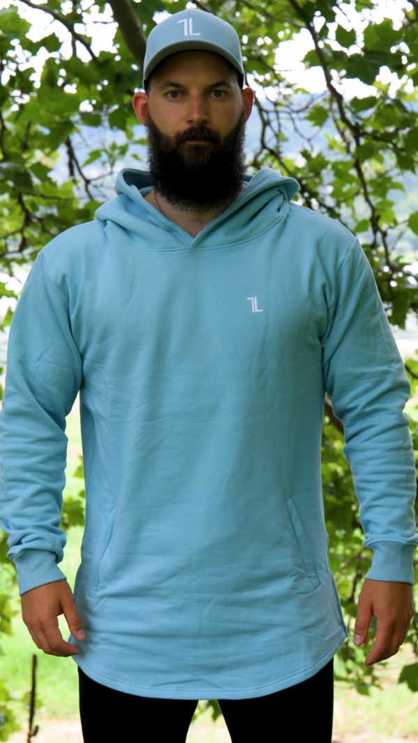 A bearded man stands outdoors before leafy trees, wearing a SPEKTR Hoodie in sky blue for high performance and a matching cap, both featuring a small white logo on the chest and hat.