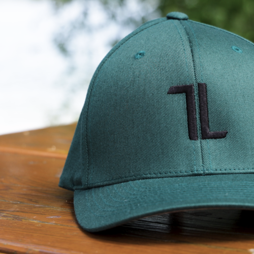The Original Ball Cap - Spruce, featuring tall panels and a black "L" logo on the front, sits on a wooden outdoor table with blurred greenery in the background.