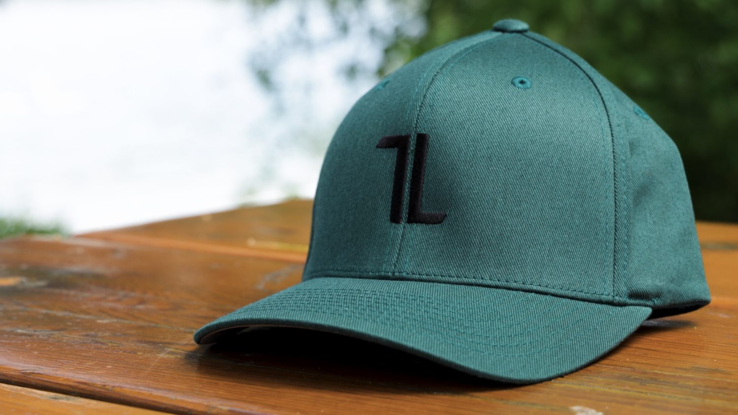 A spruce Original Ball Cap with a black “1L” logo and tall panels rests on a wooden surface outdoors, T∙LIV crest visible, against blurred greenery.