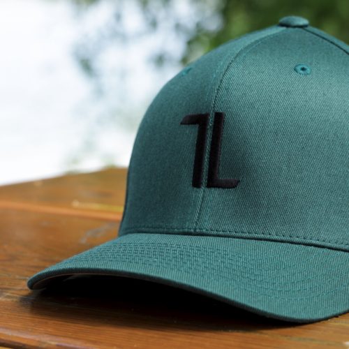 A spruce Original Ball Cap with a black “1L” logo and tall panels rests on a wooden surface outdoors, T∙LIV crest visible, against blurred greenery.