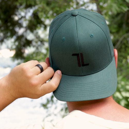 Someone wearing the Original Ball Cap - Spruce with T∙LIV crest and tall panels stands outdoors, holding the brim. Trees and greenery create a blurred background.
