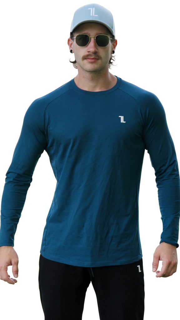 A man stands facing forward, wearing the FRMR Long sleeve - Deep Ops gym shirt in blue with a small white "7L" logo, black pants, a light gray cap also featuring the white "7L" logo, and sunglasses.