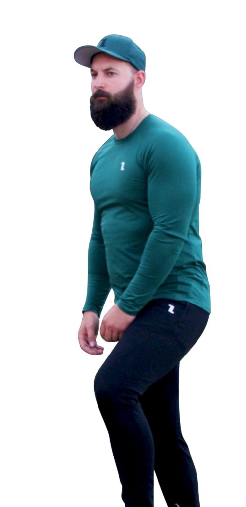 A bearded man in a FRMR Long Sleeve - Recon Pine shirt, black pants, and a green cap stands against a white background, slightly bent forward with one arm extended and a neutral expression.