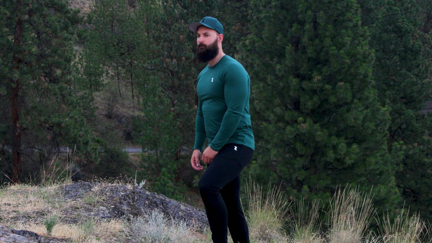 A man with a beard, wearing a green long-sleeve shirt, black pants, and a green cap, stands outdoors on rocky ground with trees in the background.