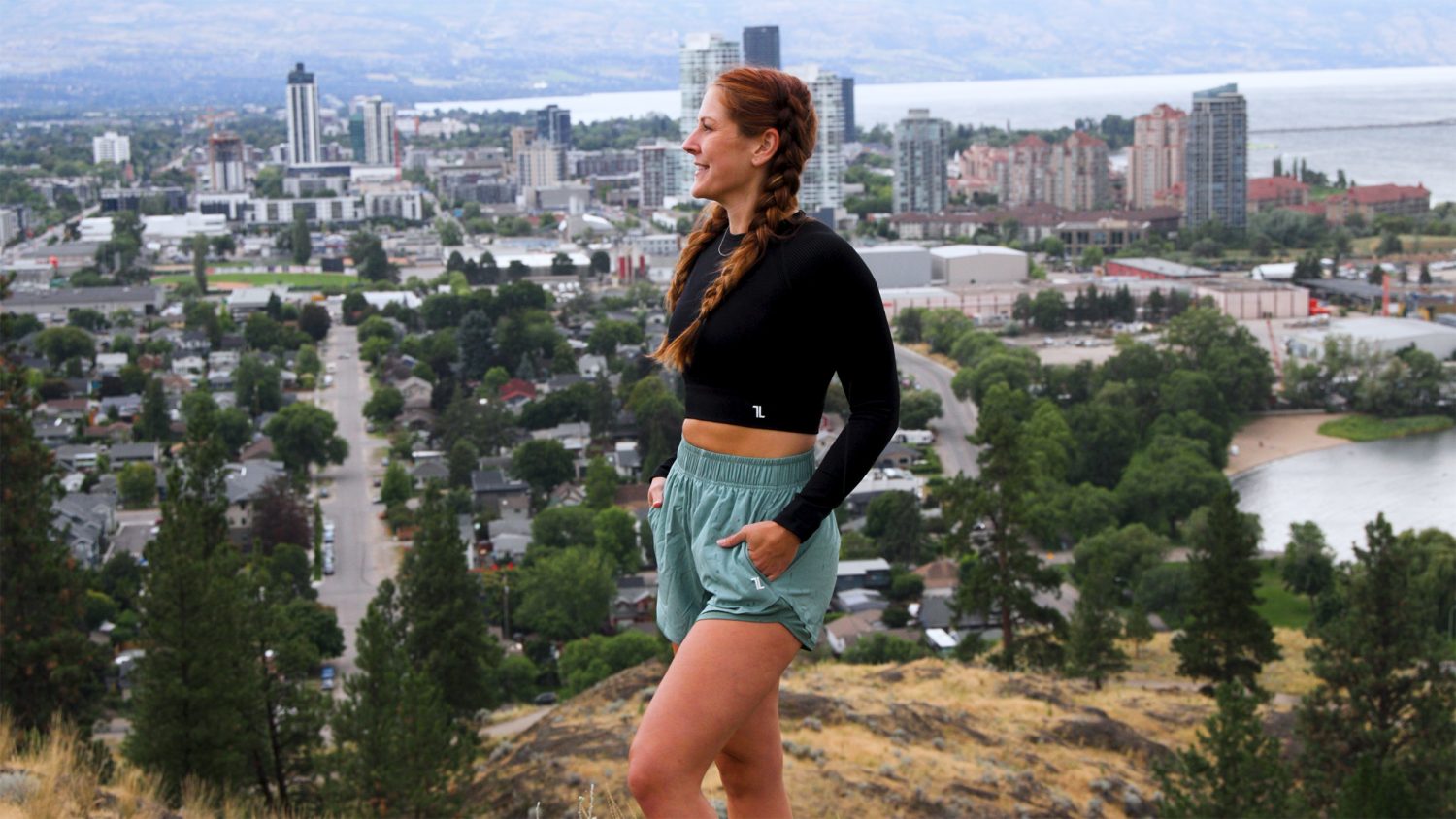 A woman with braided hair wearing a black top and green shorts stands on a hill overlooking a cityscape with buildings, trees, and a body of water in the background.