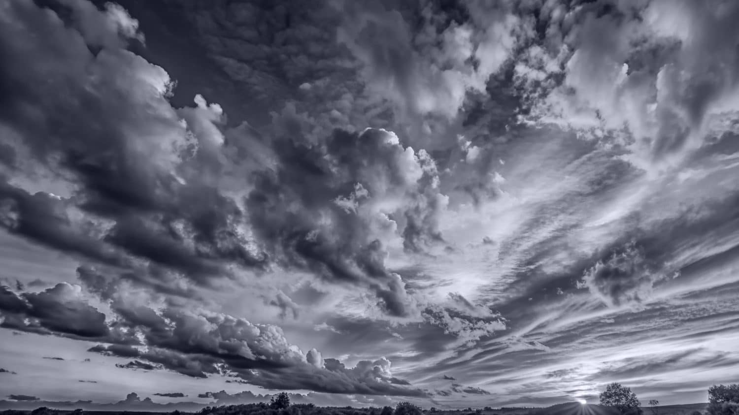 Dramatic black and white photo of a sky filled with large, billowing clouds at sunset, with the sun partially visible near the horizon and silhouettes of trees along the landscape.