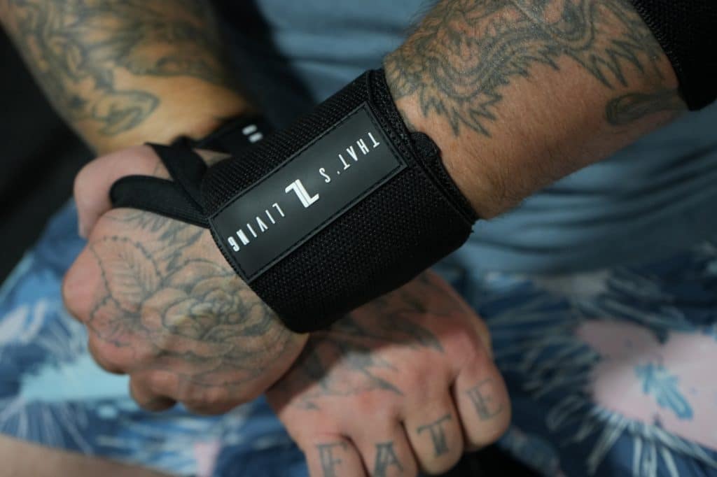 A close-up of tattooed hands, one wearing a Copy of IronHide Lifting Strap. "FATE" is tattooed on the fingers, and the person is dressed in blue and pink patterned shorts.