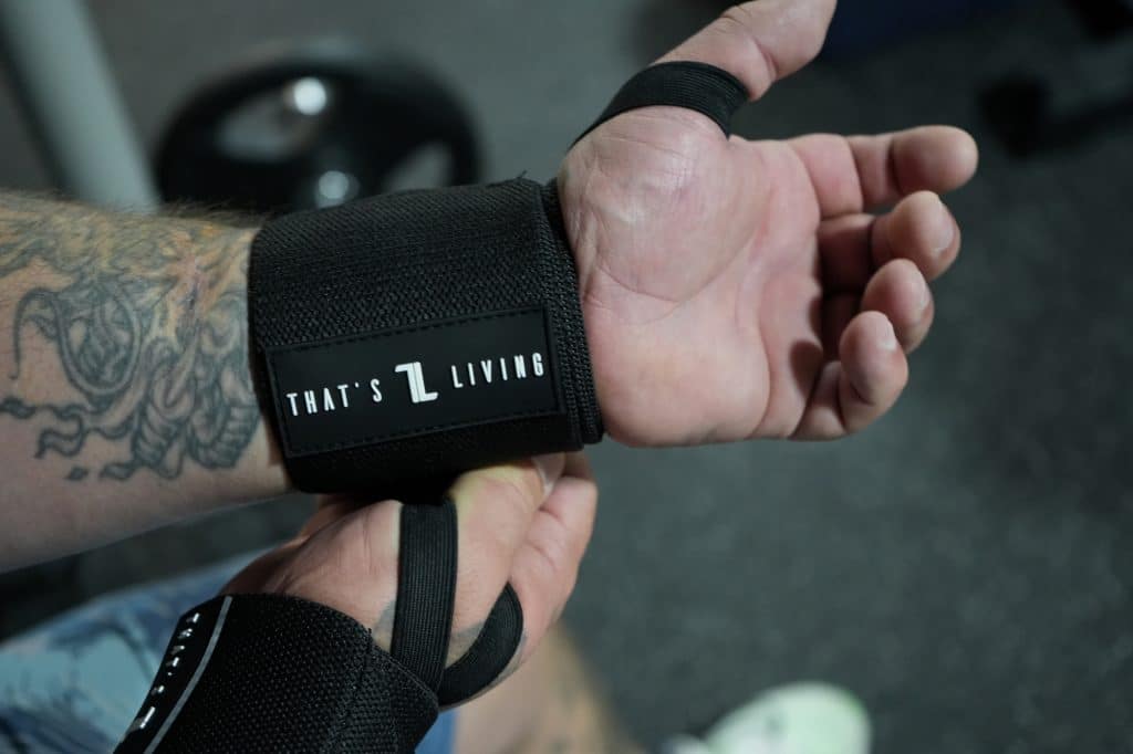A tattooed individual puts on Copy of IronHide Lifting Straps labeled "THAT'S LIVING" in a gym, with workout equipment visible in the background.