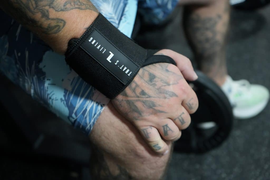 A tattooed hand with "FATE" inked across the fingers grips a weight plate, wearing Copy of IronHide Lifting Straps. The person is dressed in blue patterned shorts and white sneakers.