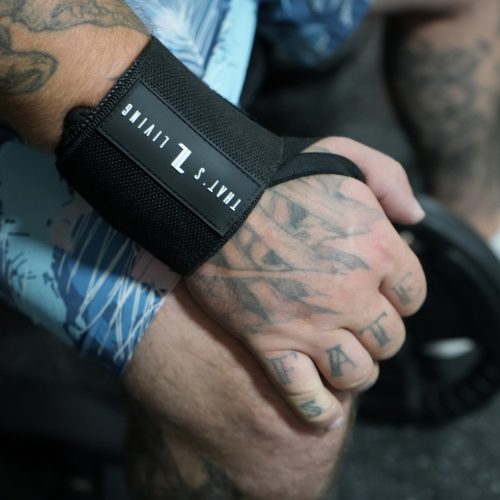 A tattooed hand with "FATE" inked across the fingers grips a weight plate, wearing Copy of IronHide Lifting Straps. The person is dressed in blue patterned shorts and white sneakers.