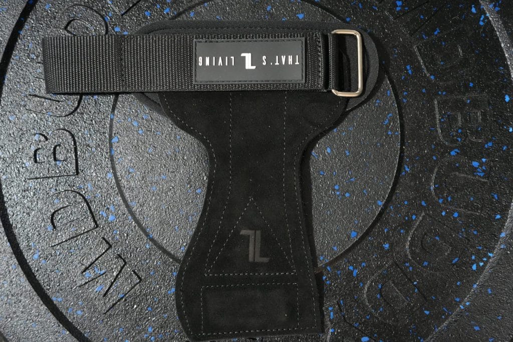 An IronHide Lifting Strap with "THAT'S LIVING" branding rests on a textured black weight plate with blue speckles and embossed markings.