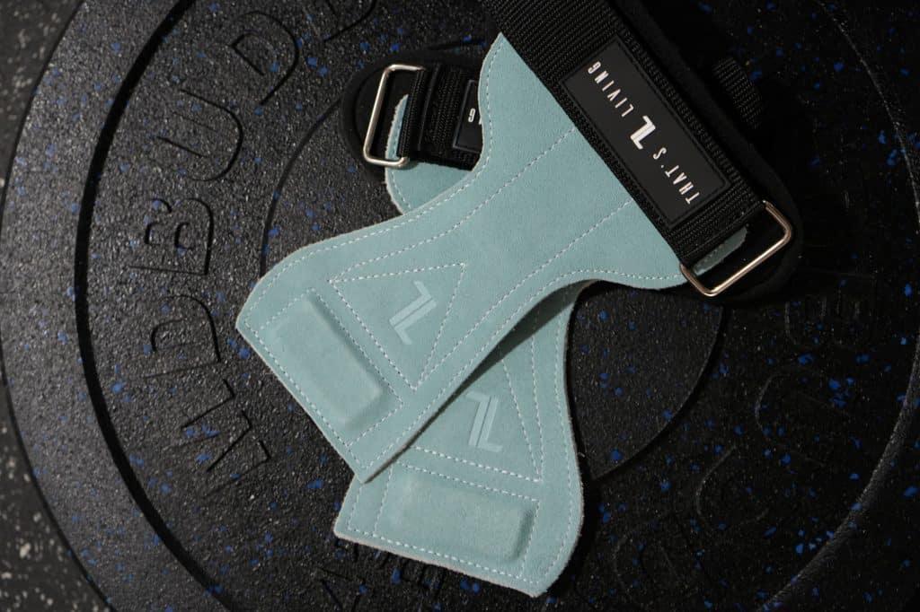 A pair of IronHide Lifting Straps in light blue suede with black straps and metal buckles rests on a black rubber weight plate embossed with text.