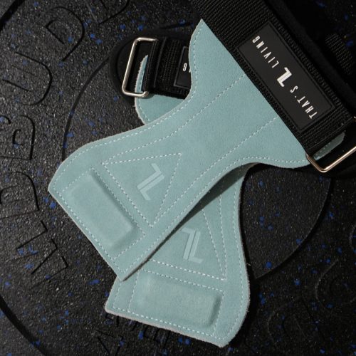 A pair of IronHide Lifting Straps in light blue suede with black straps and metal buckles rests on a black rubber weight plate embossed with text.