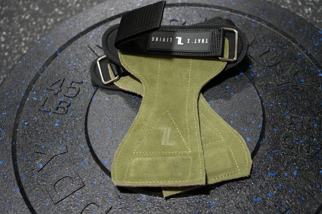 IronHide Lifting Straps in green suede with black straps are displayed on a large, black textured rubber weight plate featuring blue specks, set on a gym floor.