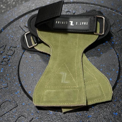 IronHide Lifting Straps in green suede with black straps are displayed on a large, black textured rubber weight plate featuring blue specks, set on a gym floor.