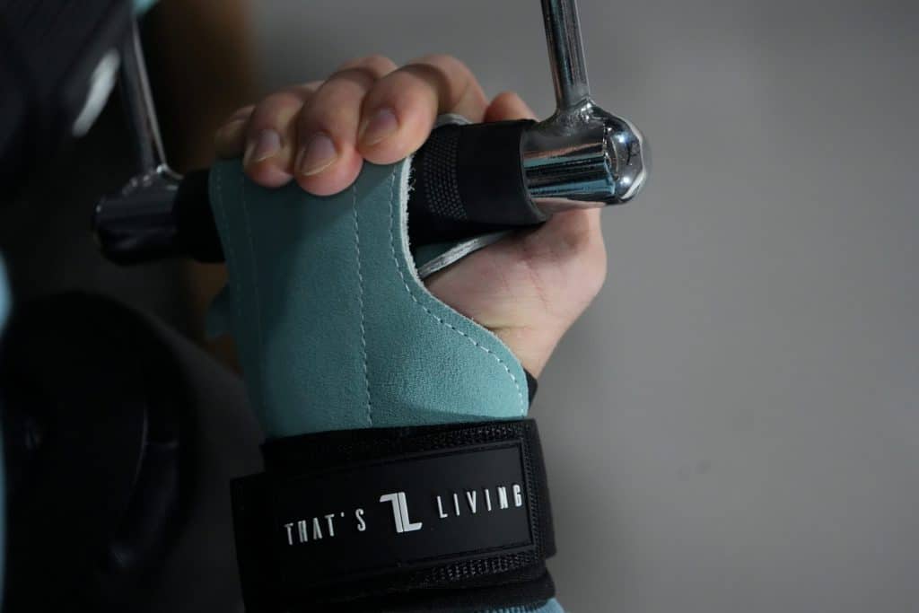 A person grips a metal gym handle using IronHide Lifting Straps, featuring a teal design with a black strap labeled "THAT'S LIVING," while the background remains blurred.