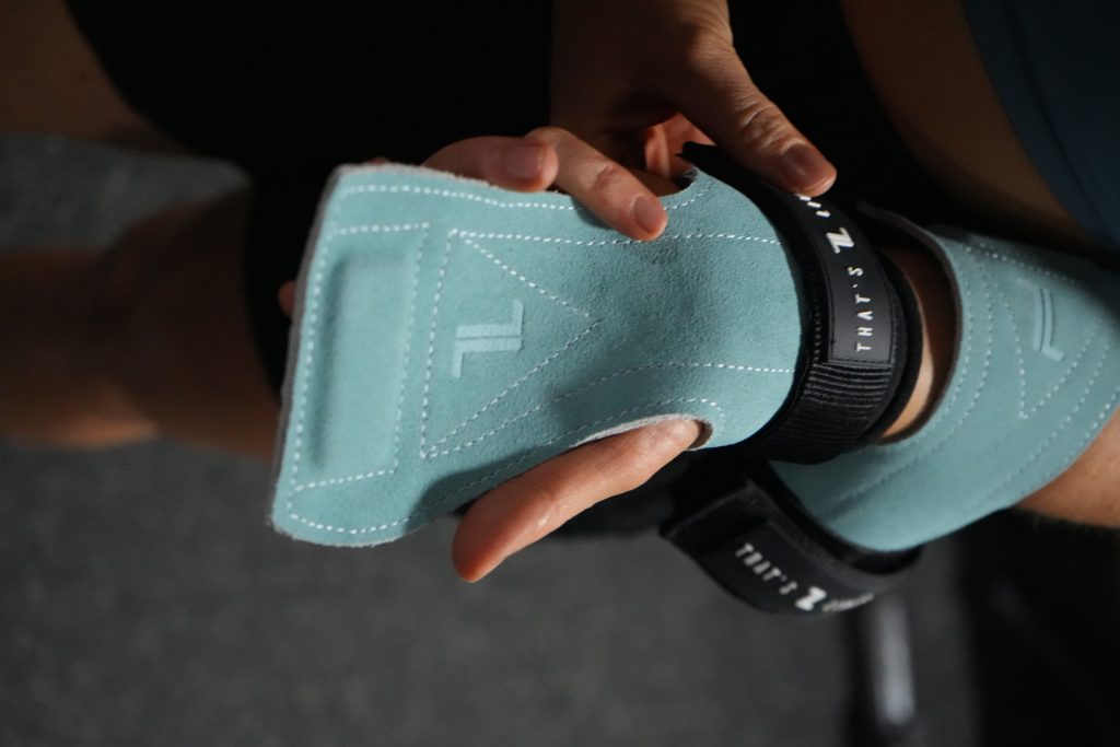 A hand wears IronHide Lifting Straps in light blue with black straps and white stitching, featuring a stylized "TL" logo. The person prepares for a workout, while the background is out of focus.