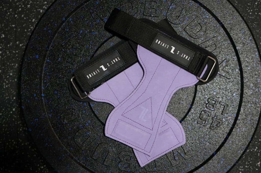 A pair of IronHide Lifting Straps – Black with white branding rests on a black 45-pound rubber-coated weight plate. The background is a speckled gym floor.