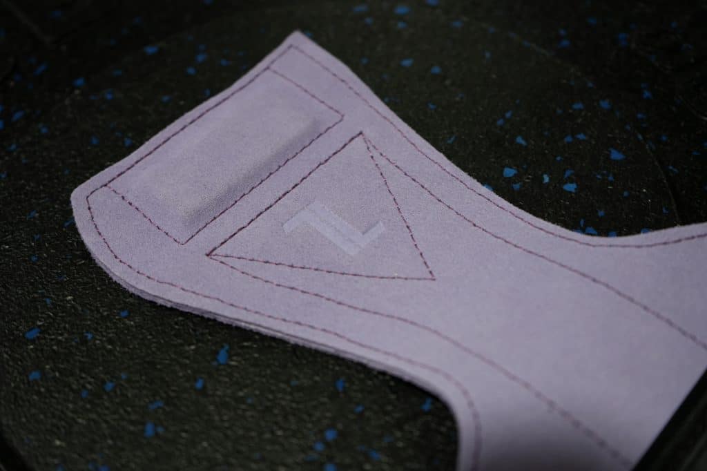 A close-up of the IronHide Lifting Straps – Black shows their suede-like purple fabric patch with visible seams and a subtle "Z" logo, resting on a black speckled surface.