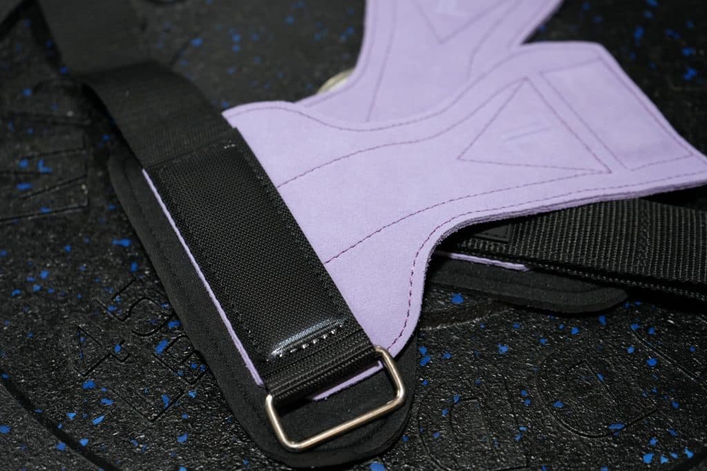 Close-up of IronHide Lifting Straps – Black, featuring a metal buckle, lying on a textured black gym mat with blue specks.