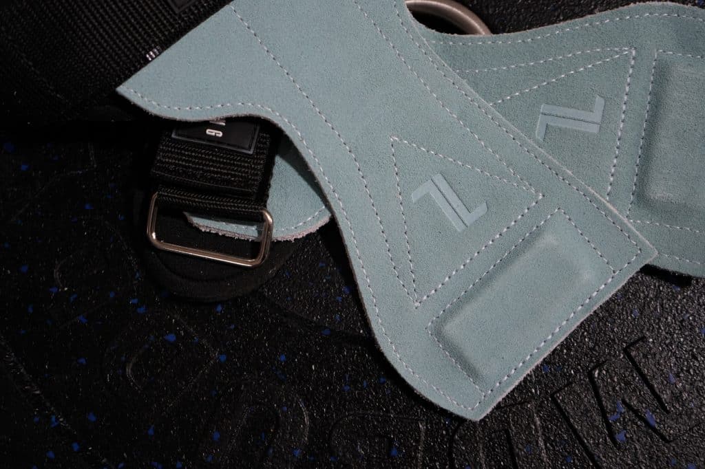 A close-up of IronHide Lifting Straps in teal suede with stitched designs and metal D-rings, displayed on a black gym mat featuring subtle blue speckles.