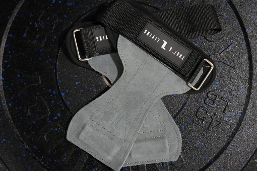 A pair of IronHide Lifting Straps with gray suede grips, black straps, and metal loops rest crossed on a black 45 lb weight plate with blue specks.