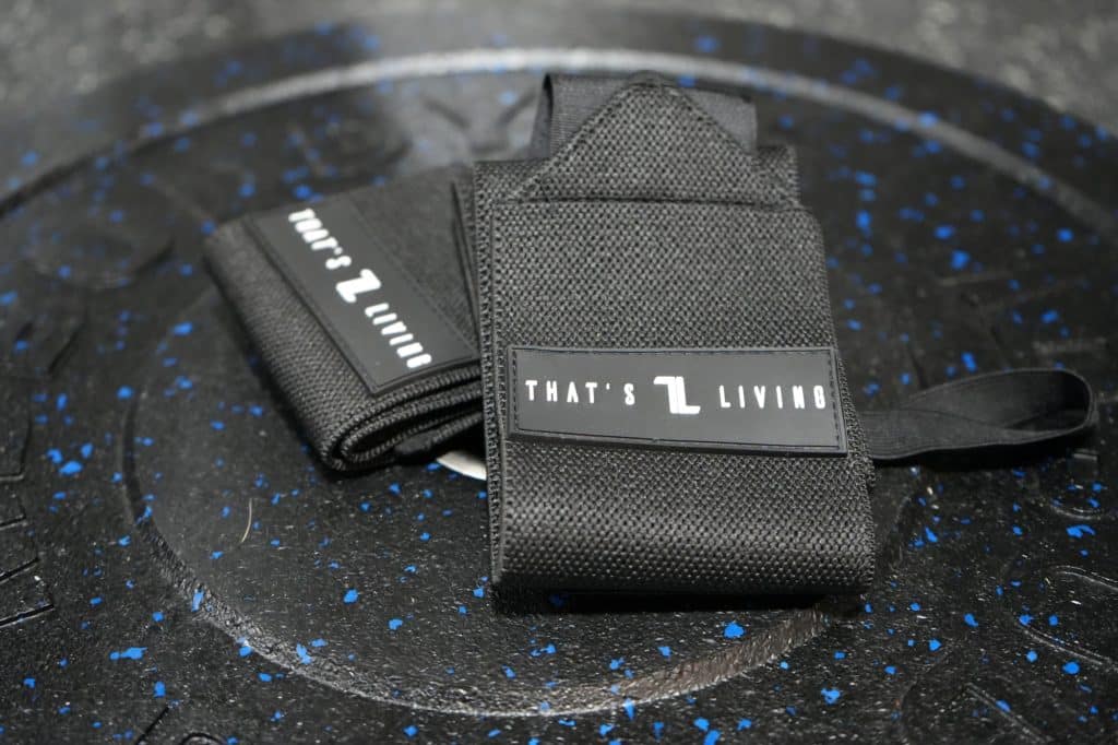 A pair of black Copy of IronHide Lifting Straps with "THAT'S LIVING" text rests on a speckled black gym weight plate.