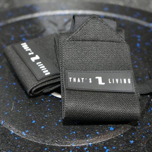 A pair of black Copy of IronHide Lifting Straps with "THAT'S LIVING" text rests on a speckled black gym weight plate.