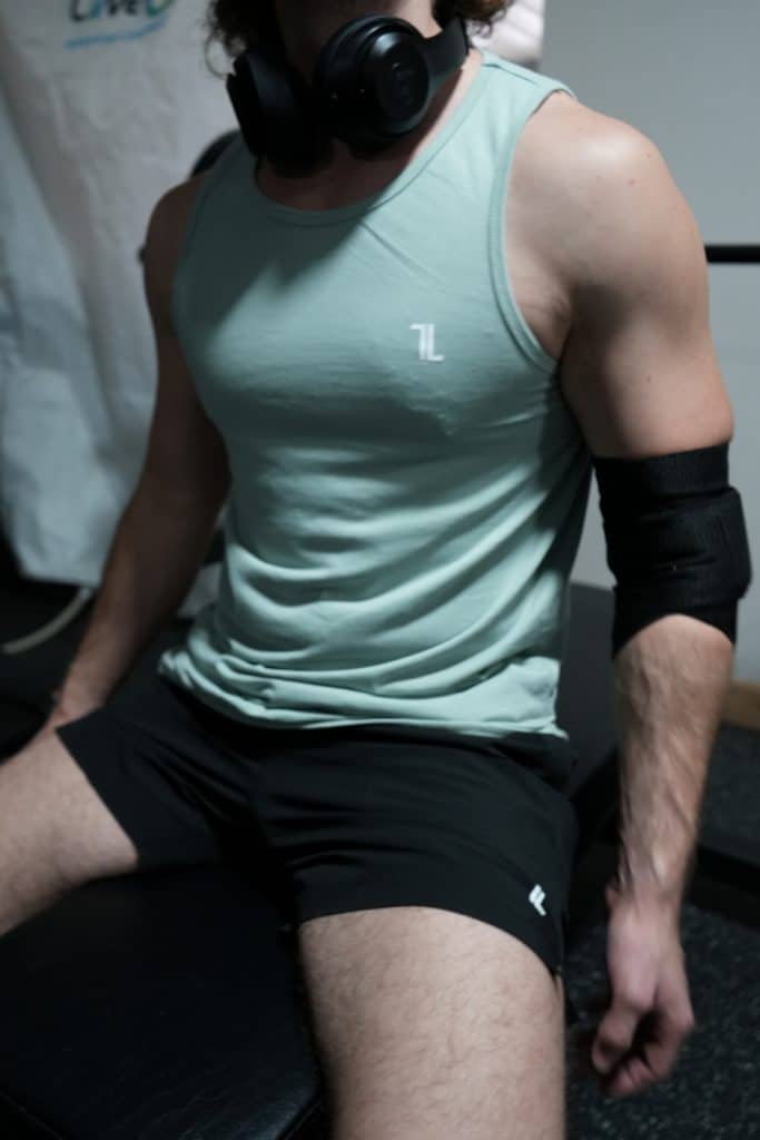 A person wearing a mint green tank top, black shorts, black headphones, and the Original Elbow Wraps sits on a bench in a gym.