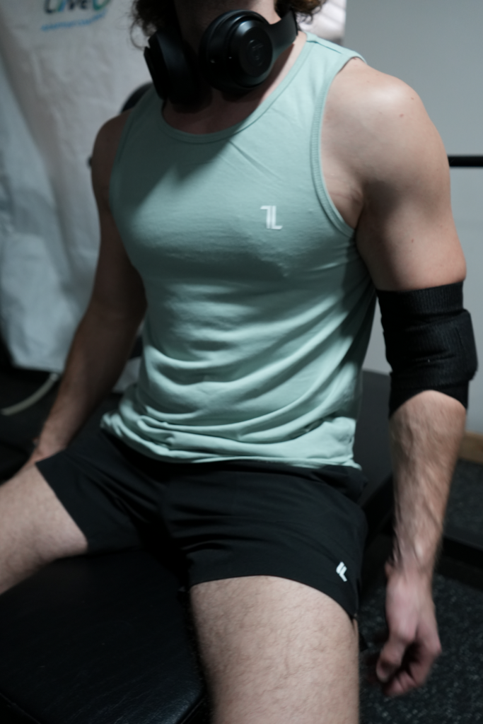 A person wearing Original Elbow Wraps, a mint green tank top, black shorts, and over-ear headphones sits on a bench in a gym setting.
