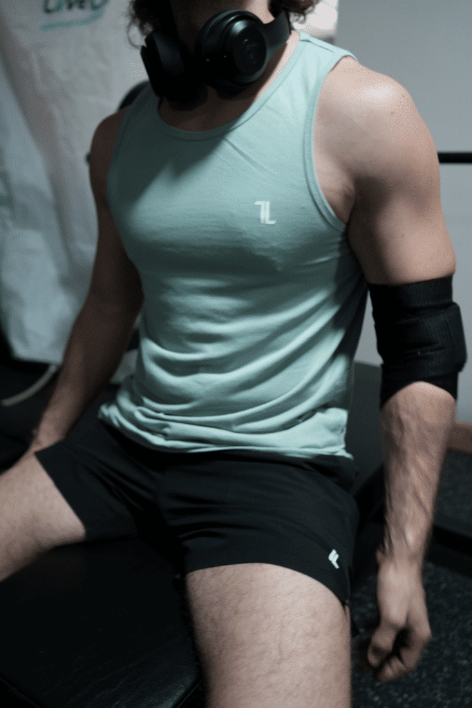 A person wearing Original Elbow Wraps, a mint green tank top, black shorts, and over-ear headphones sits on a bench in a gym setting.