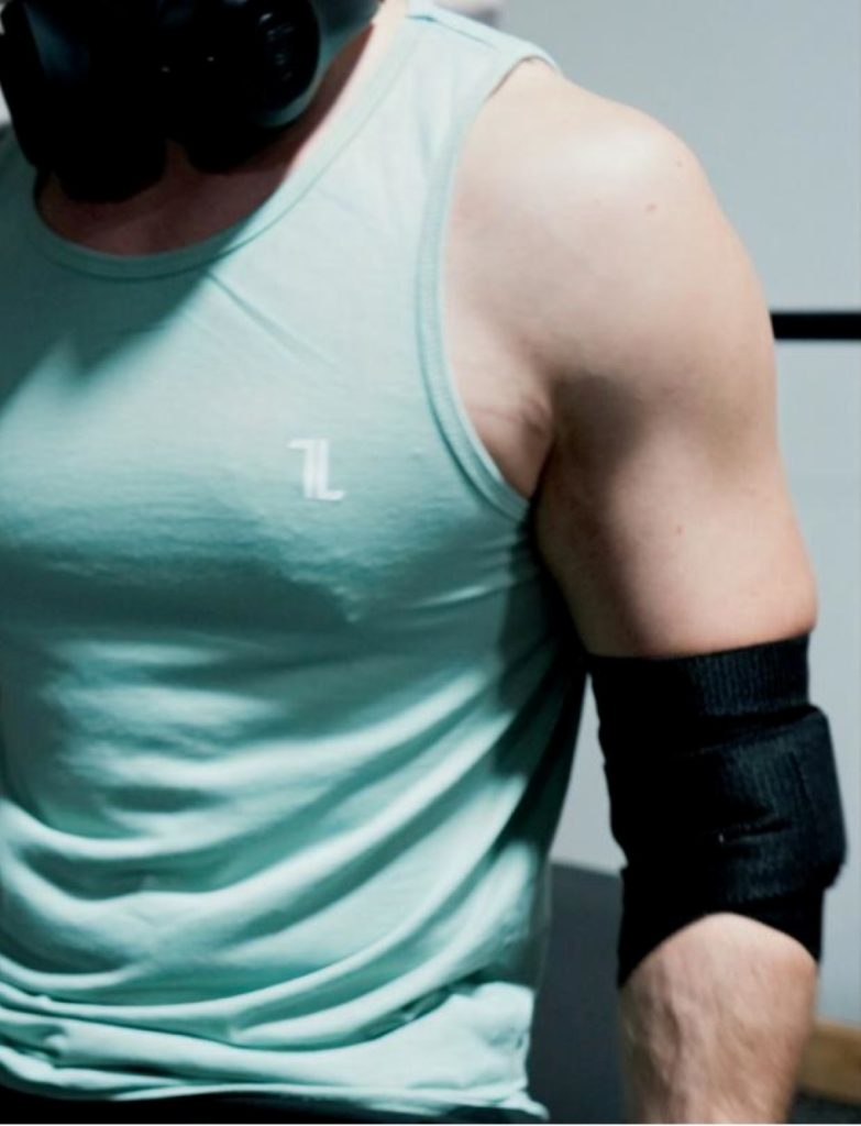 A person at the gym, shown from shoulders to waist, wears a light blue tank top, black headphones, and Original Elbow Wraps.