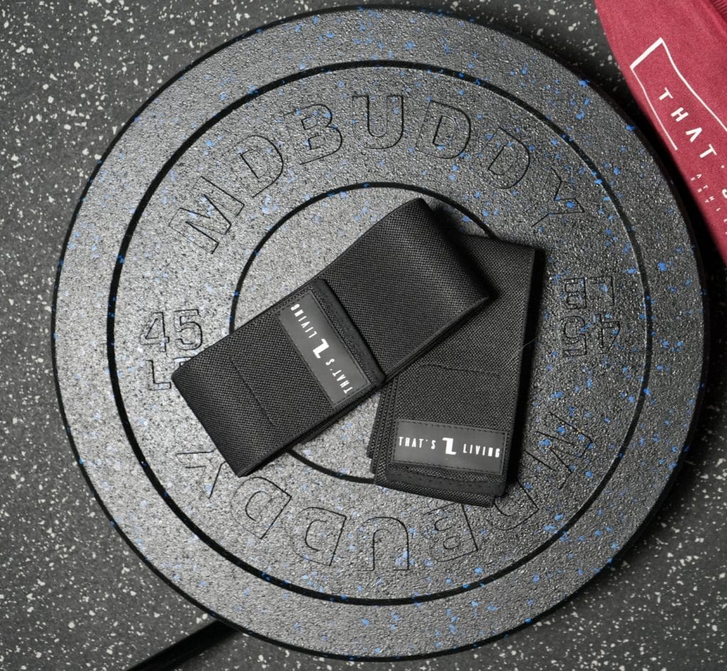 Two black Original Elbow Wraps labeled "THAT'S L LIVING" are placed on top of two stacked black MD Buddy 45-pound weight plates, set on a speckled gym floor. A portion of a red cloth appears in the corner.
