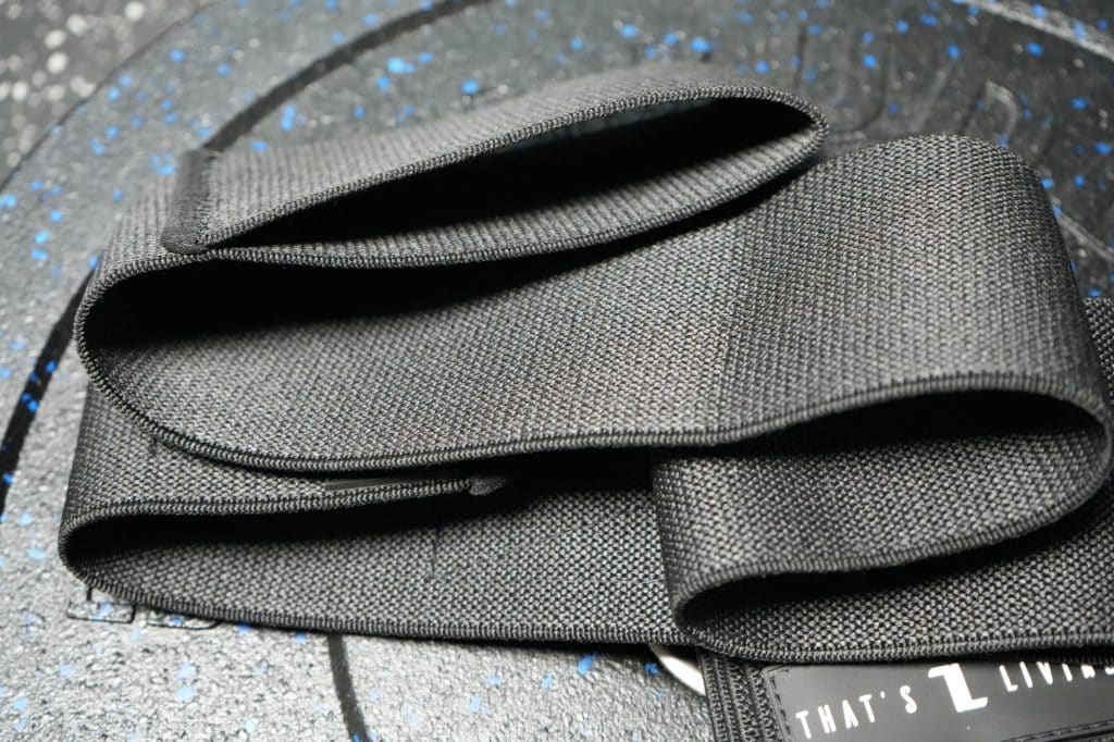 A close-up of an Original Elbow Wrap, black fabric, folded on a textured dark surface with blue speckles. A label with partial text appears in the lower right corner.
