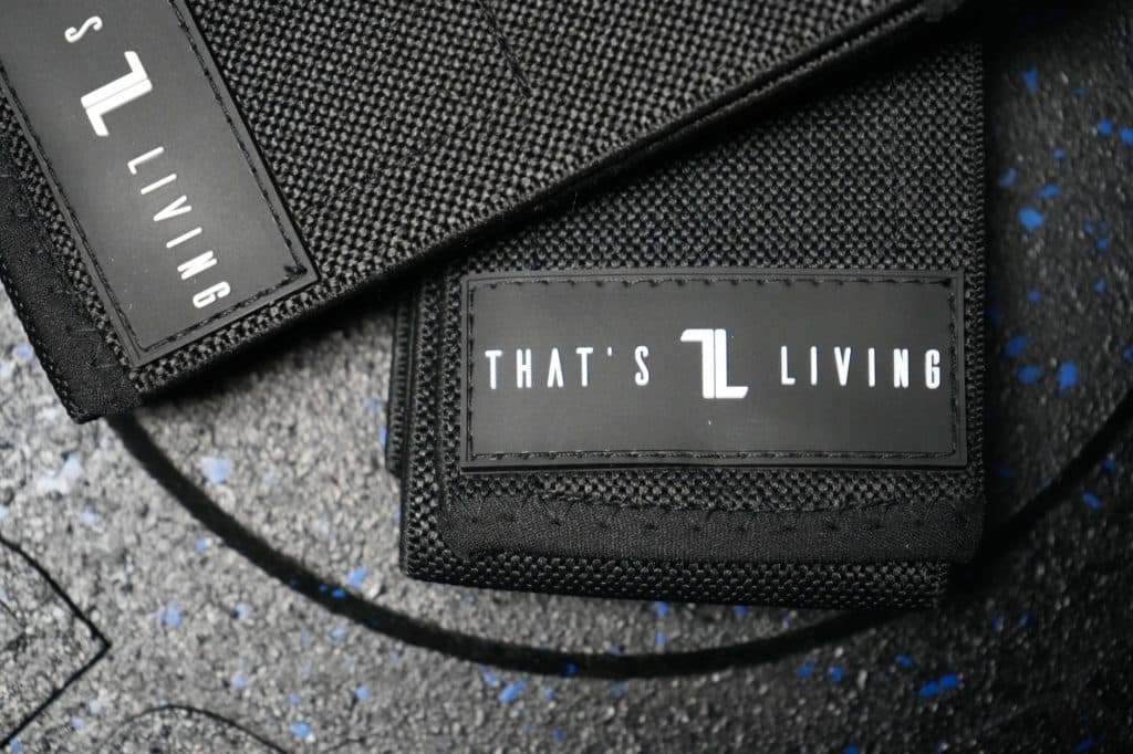 Close-up of two black Original Elbow Wraps with "THAT'S LIVING" labels in white text, placed on a textured black surface featuring blue specks.
