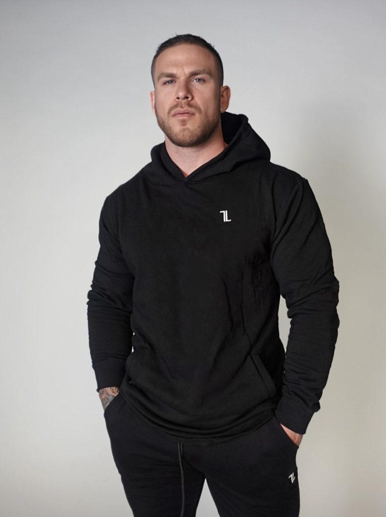 A man with short hair and a trimmed beard stands against a plain light background, wearing a black hoodie and matching black pants, with his hands in his pockets.