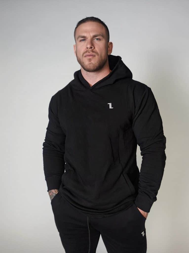 A man with short hair and a trimmed beard stands against a plain light background, wearing a black hoodie and matching black pants, with his hands in his pockets.