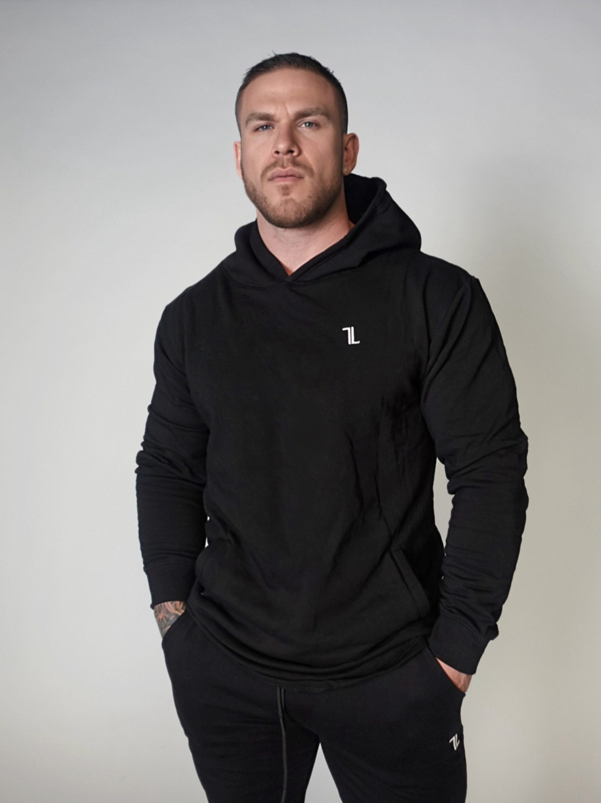 A man with short hair and a trimmed beard stands against a plain light background, wearing a black hoodie and matching black pants, with his hands in his pockets.
