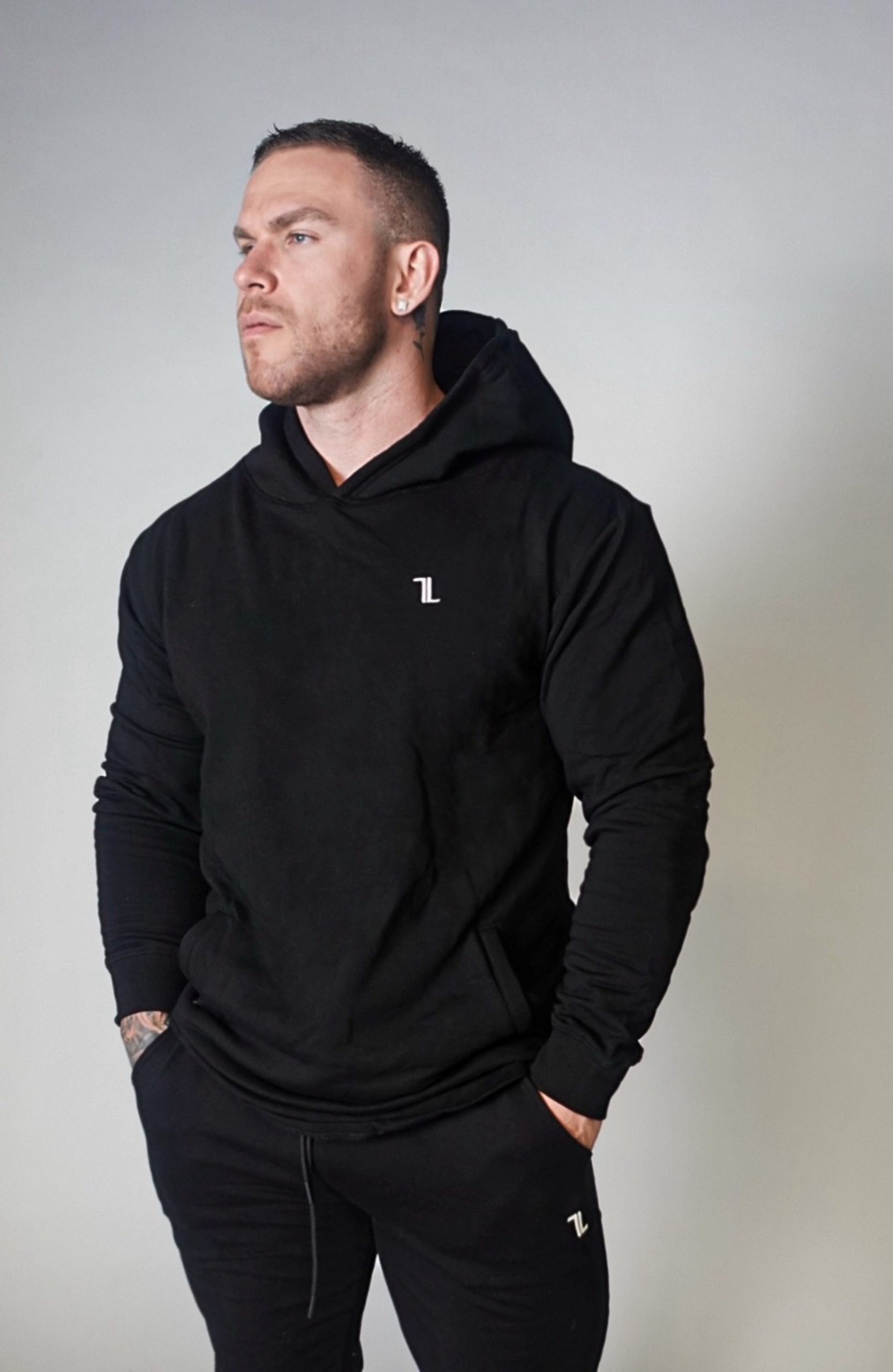 A man stands against a plain light background wearing the SPEKTR Hoodie—black with a small white logo on the chest—and matching black pants, hands in his pockets, looking off to the side.