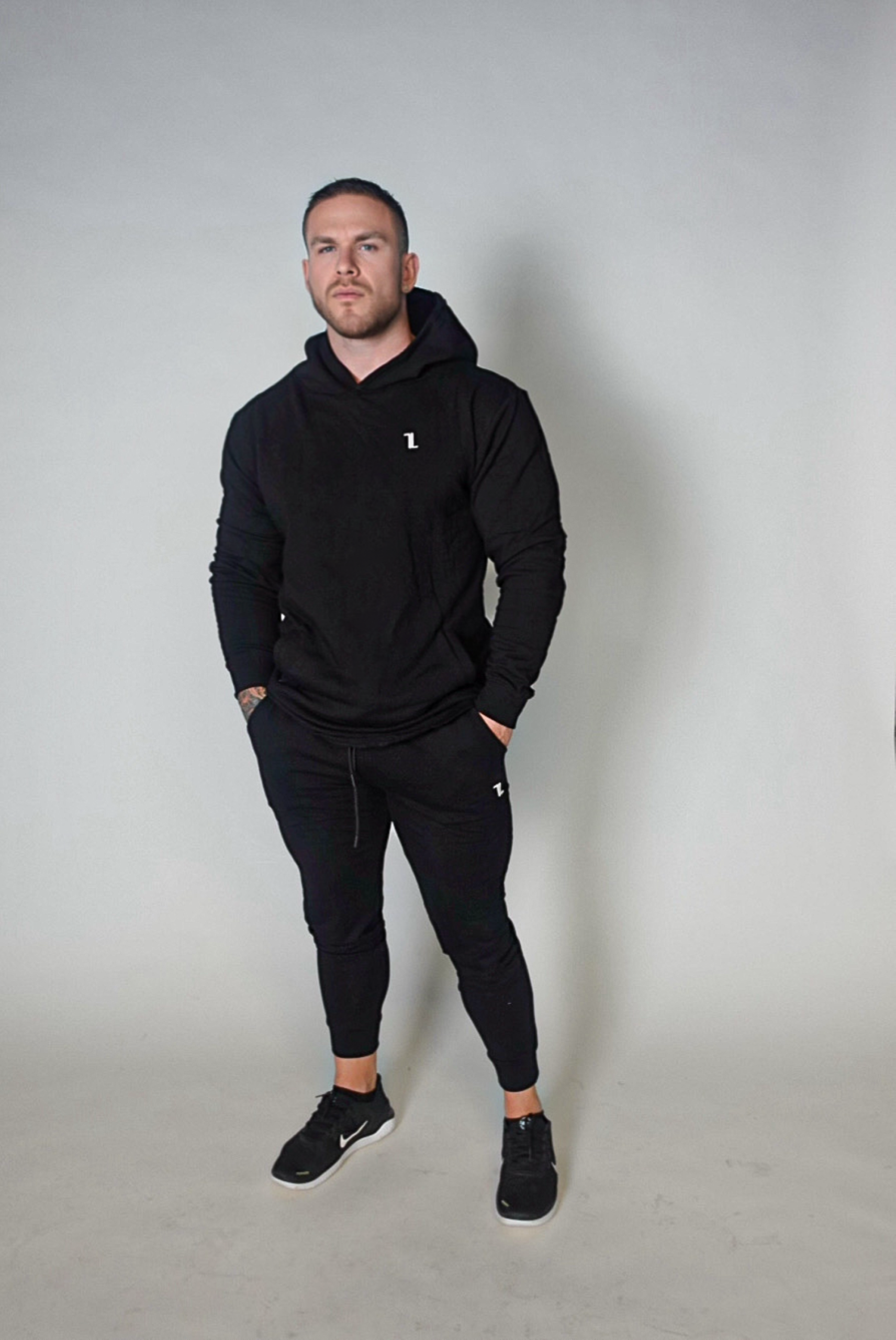 A man stands facing forward against a plain light background, wearing the black SPEKTR Hoodie, black jogger pants, and black sneakers. His hands are in his pockets and he has a neutral expression.