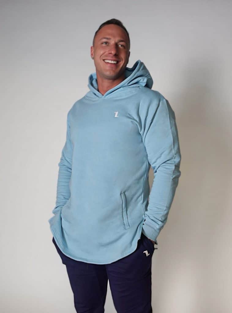 A man smiles and looks to the side with his hands in the pockets of a light blue SPEKTR Hoodie, standing against a plain light background.