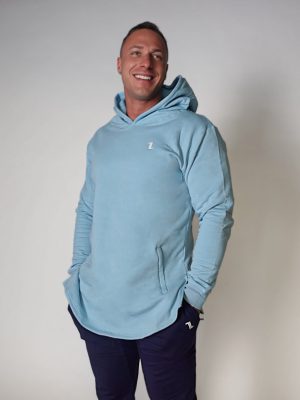 A smiling man stands against a plain background, wearing a light blue hooded sweatshirt with a small "Z" logo and dark pants. His hands are in his pockets and the hood is up on his head.