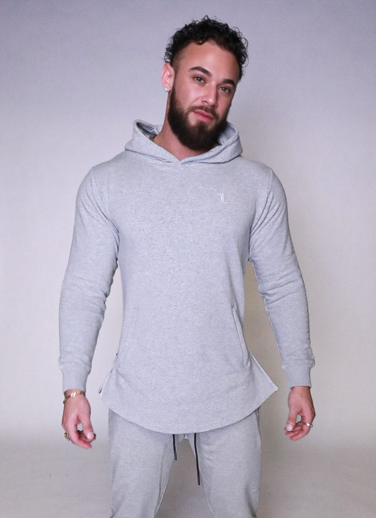 A man with a beard and curly hair wears a light gray hooded sweatshirt and matching sweatpants, standing against a plain light background, looking at the camera with a relaxed expression.