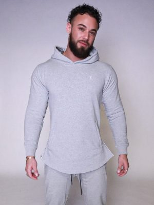 A man with a beard and curly hair wears a light gray hooded sweatshirt and matching sweatpants, standing against a plain light background, looking at the camera with a relaxed expression.