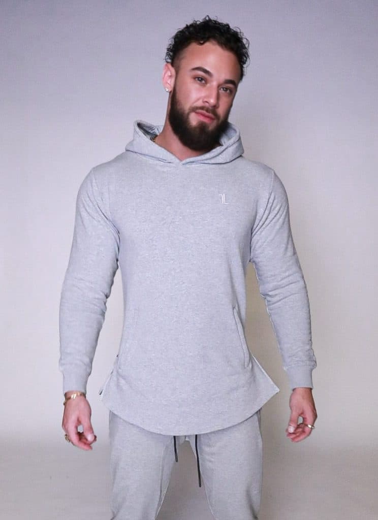 A man with a beard and curly hair wears a light gray hooded sweatshirt and matching sweatpants, standing against a plain light background, looking at the camera with a relaxed expression.