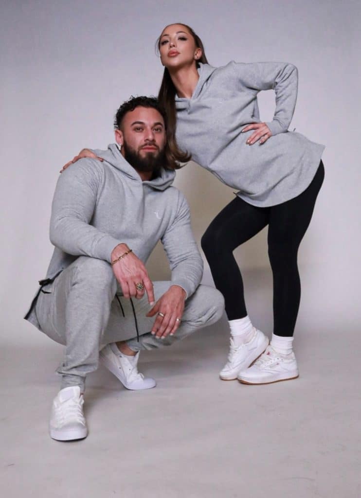 A man squats while a woman stands beside him, both confidently posing in matching SPEKTR Hoodies with black leggings and white sneakers against a plain light background.