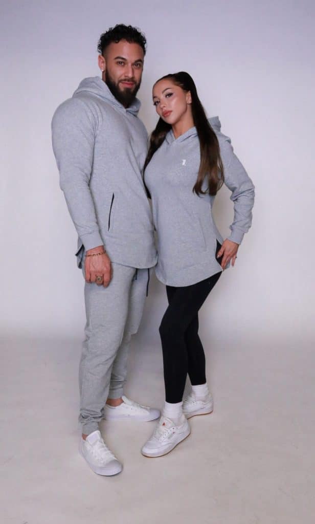 A man and woman stand close against a plain background, both confidently facing the camera while wearing matching light gray SPEKTR Hoodies, black pants or leggings, and white sneakers.