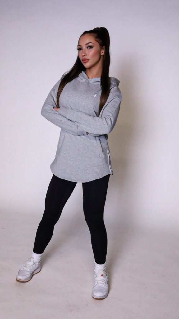 A woman stands confidently with arms crossed, wearing the gray SPEKTR Hoodie, black leggings, and white sneakers against a plain white background.