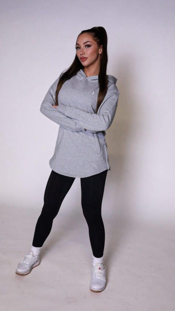 A woman stands confidently with arms crossed, wearing the gray SPEKTR Hoodie, black leggings, and white sneakers against a plain white background.