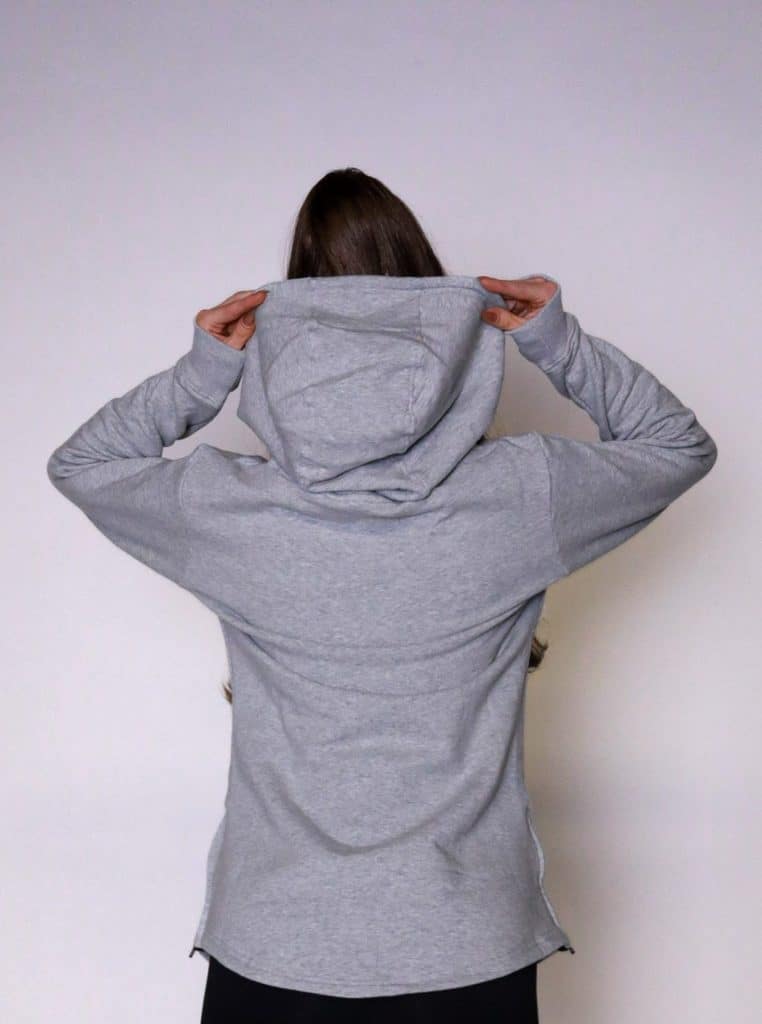 A person with long brown hair, seen from behind, wears the SPEKTR Hoodie in light gray and lifts the hood with both hands against a plain light background.